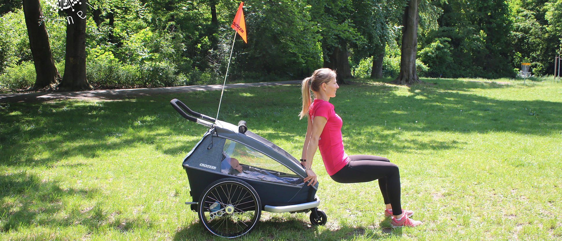 Woman performing dip exercise using Croozer bike trailer for support with child inside in sunny park